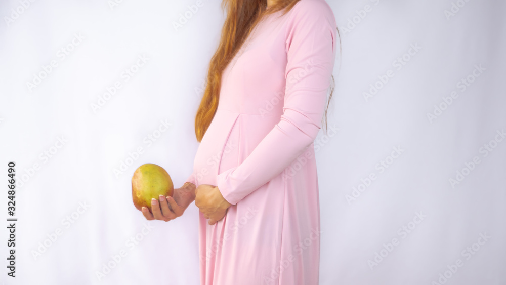 A close up photo of a pregnant young woman holding a mango next to her belly showing the size of the baby in a beautiful dress. Photos of fetal growth at 19 week pregnancy. Healthy pregnancy diet. A close up photo of a pregnant young woman holding a mango next to her belly showing the size of the baby in a beautiful dress. Photos of fetal growth at 19 week pregnancy. Healthy pregnancy diet.