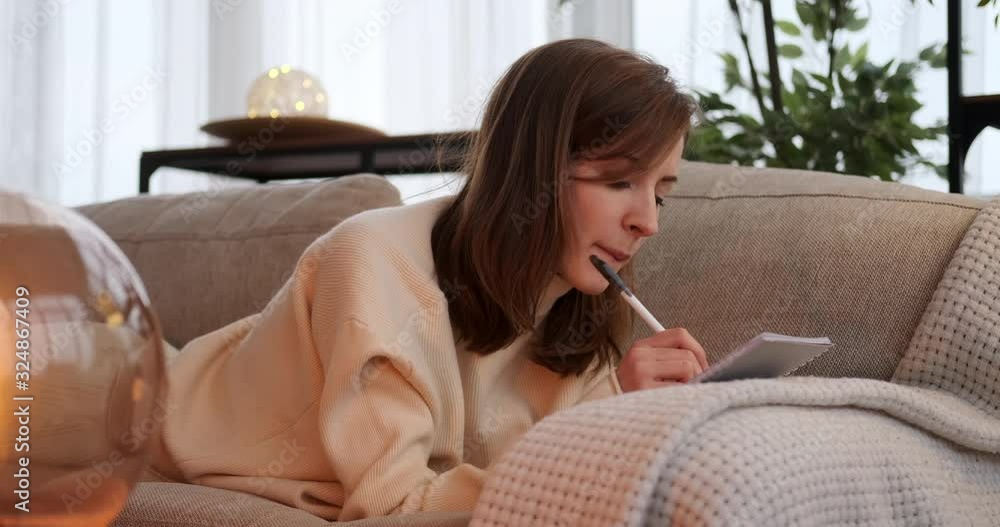 Beautiful woman thinking and writing in notepad while relaxing on sofa at home Beautiful woman thinking and writing in notepad while relaxing on sofa at home