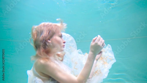 Beautiful blonde girl swims underwater in the pool in a fluffy white dress. Looks like a nymph or a mermaid