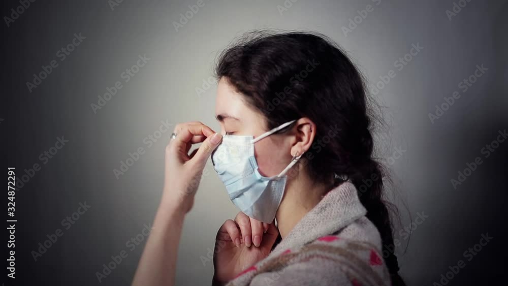 A young Caucasian woman with a sickly appearance, puts a medical mask on her face. Dark background. Side view. The concept of protection against viral infections A young Caucasian woman with a sickly appearance, puts a medical mask on her face. Dark background. Side view. The concept of protection against viral infections