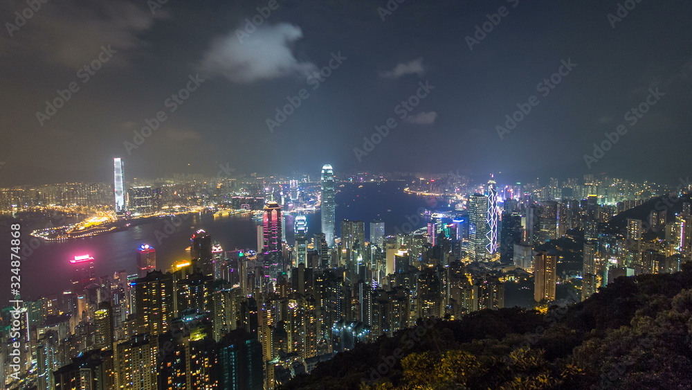 Hong Kong city skyline timelapse at night with Victoria Harbor and skyscrapers illuminated by lights over water viewed from mountain top.