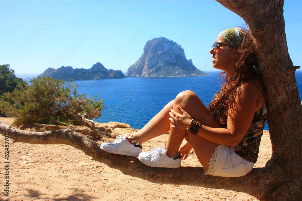 summer vacation of a brunette caucasian girl sitting on a tree branch admiring the islet of Es Vedra in Cala dhort