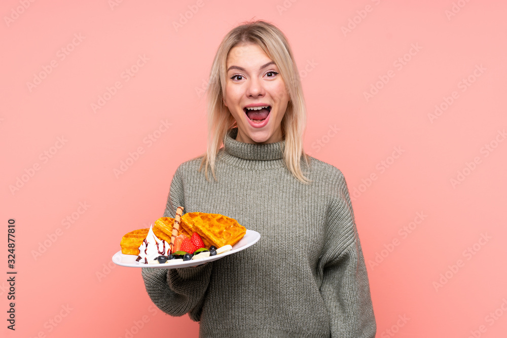 Young blonde woman holding waffles over isolated pink background with surprise and shocked facial expression Young blonde woman holding waffles over isolated pink background with surprise and shocked facial expression