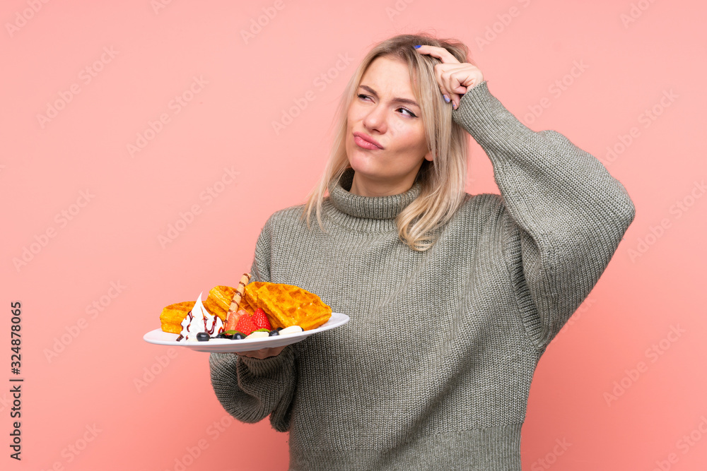 Young blonde woman holding waffles over isolated pink background having doubts and with confuse face expression Young blonde woman holding waffles over isolated pink background having doubts and with confuse face expression