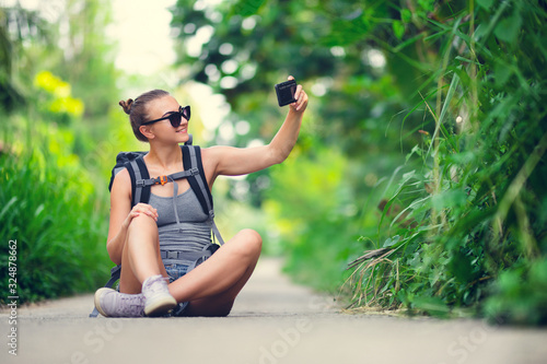 Fototapeta Naklejka Na Ścianę i Meble -  Young pretty white Caucasian girl with hiking backpack takes selfie while sitting on a jungle trail. Girl traveler with glasses make