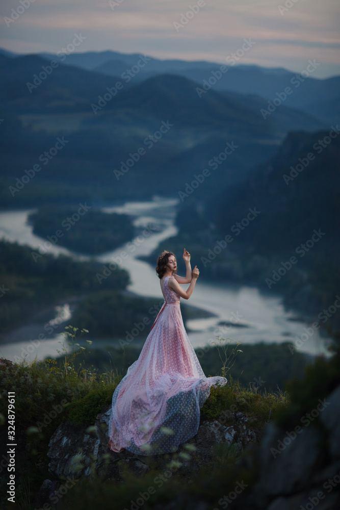 A girl in a pink dress with a long train is dancing on the top of the mountain
