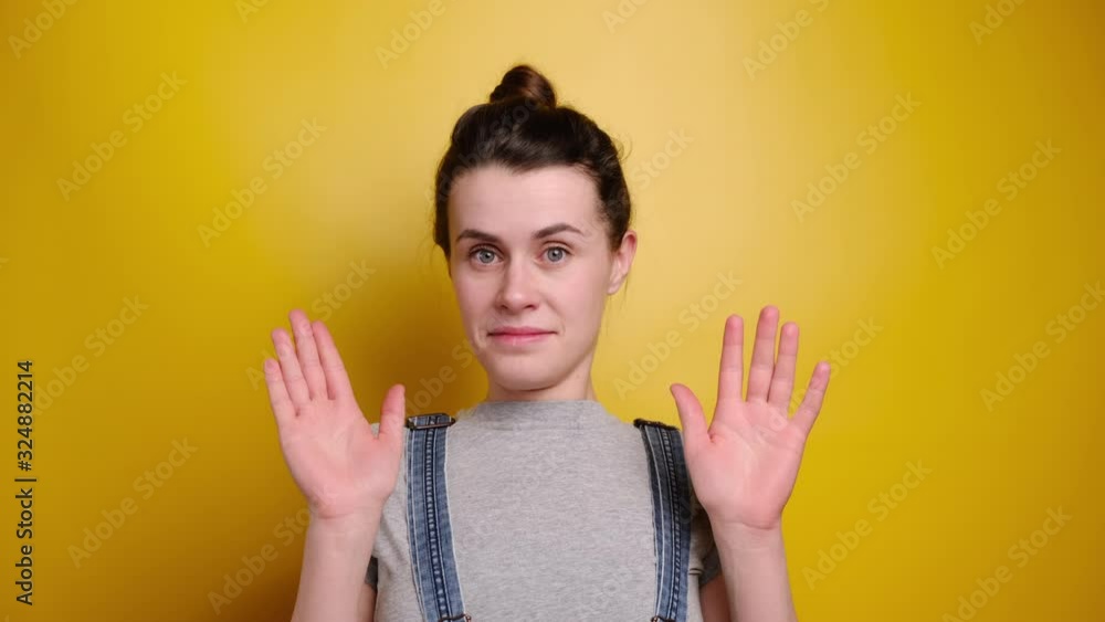 Attractive brunette female shows refusal gesture, doesn`t want to participate in meeting, leave me in piece, wears grey t-shirt and denim overalls, poses against yellow studio background Attractive brunette female shows refusal gesture, doesn`t want to participate in meeting, leave me in piece, wears grey t-shirt and denim overalls, poses against yellow studio background