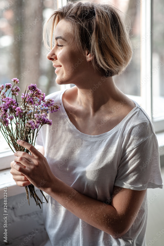 Smiling young woman near the window holds a bouquet of purple flowers near her face 