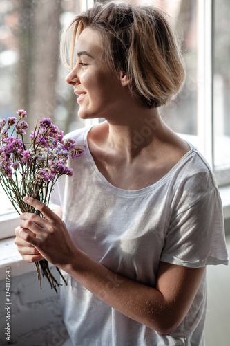 Smiling young woman near the window holds a bouquet of purple flowers near her face 