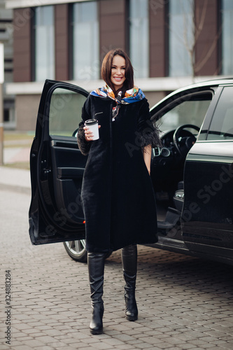 Full length of a fashionable lady in black coat and leather boots wearing colorful handkerchief around her neck. She is carrying a cup of takeaway coffee against open car in the street.