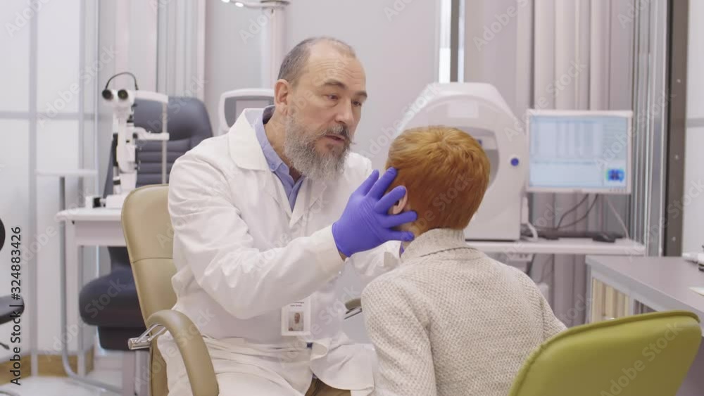 Elderly bearded ophthalmologist in lab coat and sterile gloves checking eyes of little redhead boy while working in clinic Elderly bearded ophthalmologist in lab coat and sterile gloves checking eyes of little redhead boy while working in clinic