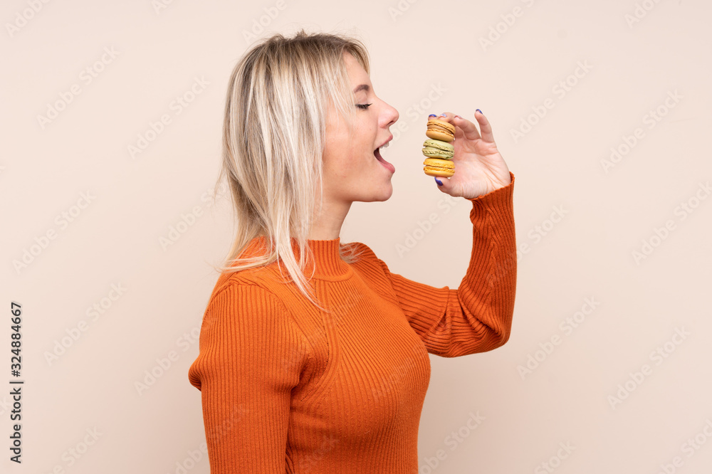 Young blonde Russian woman over isolated background holding colorful French macarons and eating it Young blonde Russian woman over isolated background holding colorful French macarons and eating it