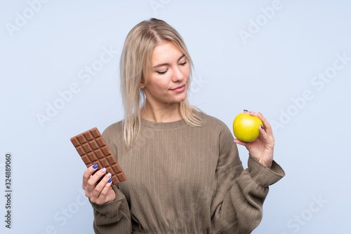 Young blonde woman over isolated blue background taking a chocolate tablet in one hand and an apple in the other