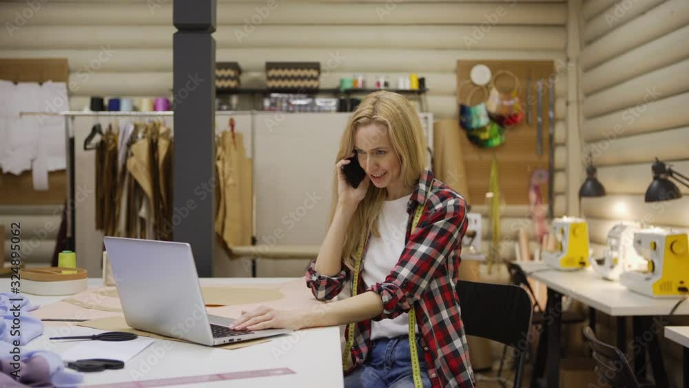 Medium shot of smiling female tailor with blond hair using laptop computer while talking on cell phone sitting at table in sewing studio Medium shot of smiling female tailor with blond hair using laptop computer while talking on cell phone sitting at table in sewing studio