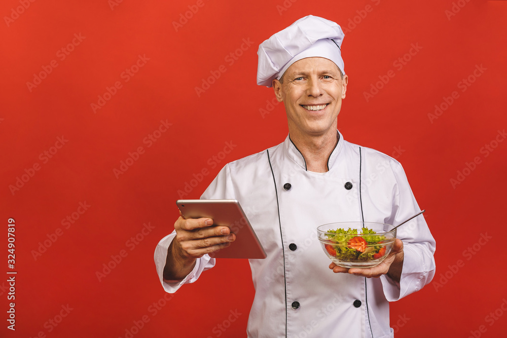 Picture of happy young senior chief cook in uniform standing isolated over red wall background, holding salad and tablet computer. Picture of happy young senior chief cook in uniform standing isolated over red wall background, holding salad and tablet computer.