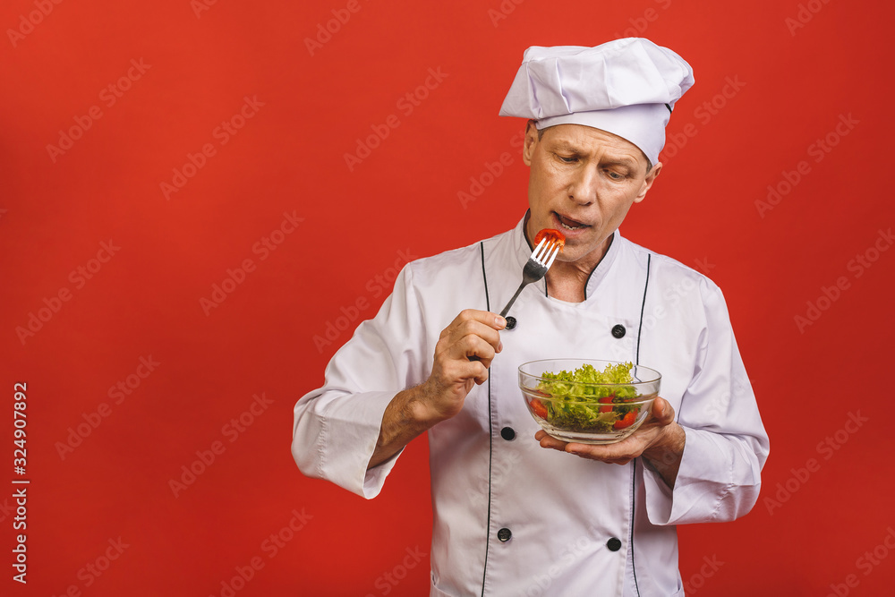 Picture of happy young senior chief cook in uniform standing isolated over red wall background, holding and eating salad. Picture of happy young senior chief cook in uniform standing isolated over red wall background, holding and eating salad.