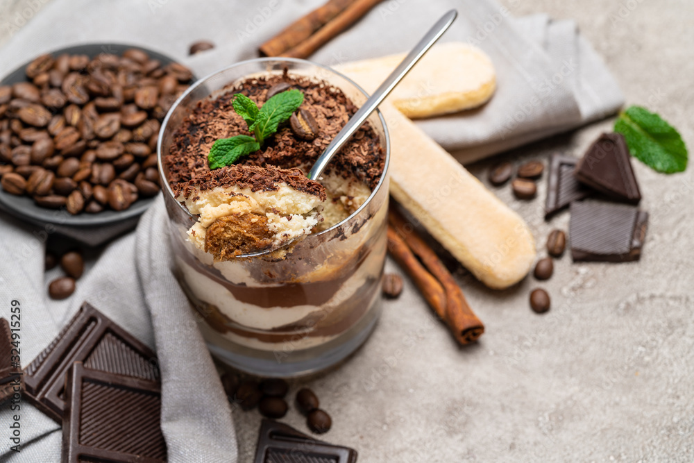 Classic tiramisu dessert in a glass cup, pieces of chocolate and savoiardi cookies on concrete background Classic tiramisu dessert in a glass cup, pieces of chocolate and savoiardi cookies on concrete background