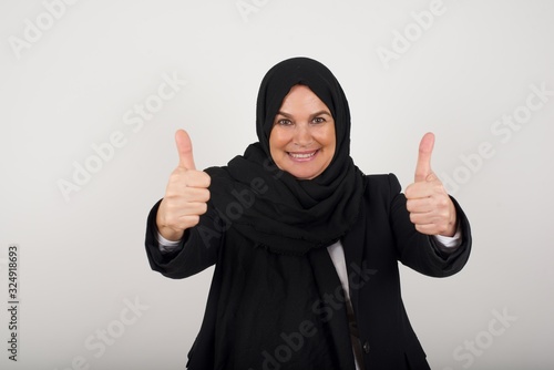 Good job! Portrait of a happy smiling muslim successful woman giving two thumbs up gesture standing indoors. Positive human emotion facial expression body language. Funny girl