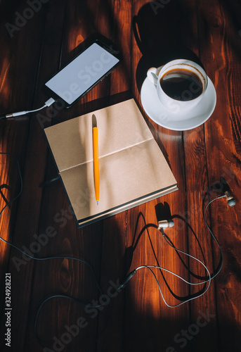 Black smartphone with white screen, paper notebook, pen, headphone and cup of coffee lies on a brown wooden background mahogany with spots of sunlight. Copy space, mockup. Music and writter concept.