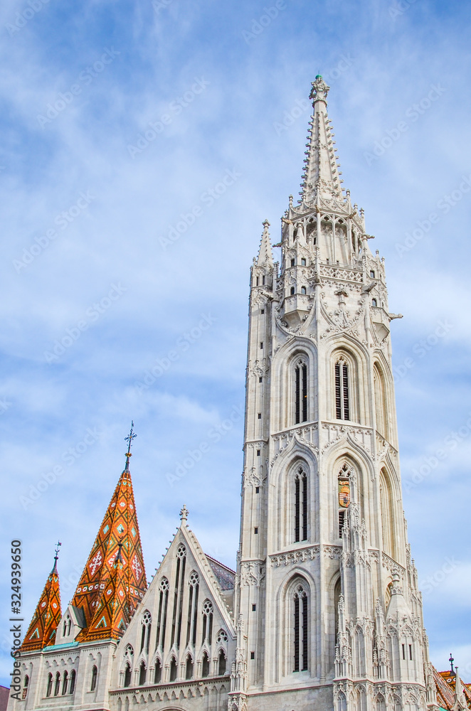 The spire of the famous Matthias Church in Budapest, Hungary. Roman Catholic church built in the Gothic style. Orange colored tile roof. Blue sky and white clouds above. Vertical photo