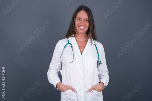 Confidence and business concept. Portrait of charming successful young doctor girl  smiling broadly with self-assured expression while holding hands in pockets standing against gray wall.