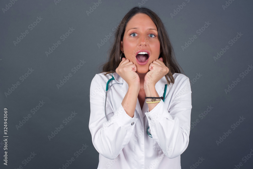 Studio shot of pleased lovely doctor woman keeps hands together under chin, being in high spirit, likes friends idea for next day, isolated on gray wall. Positive emotions. Studio shot of pleased lovely doctor woman keeps hands together under chin, being in high spirit, likes friends idea for next day, isolated on gray wall. Positive emotions.