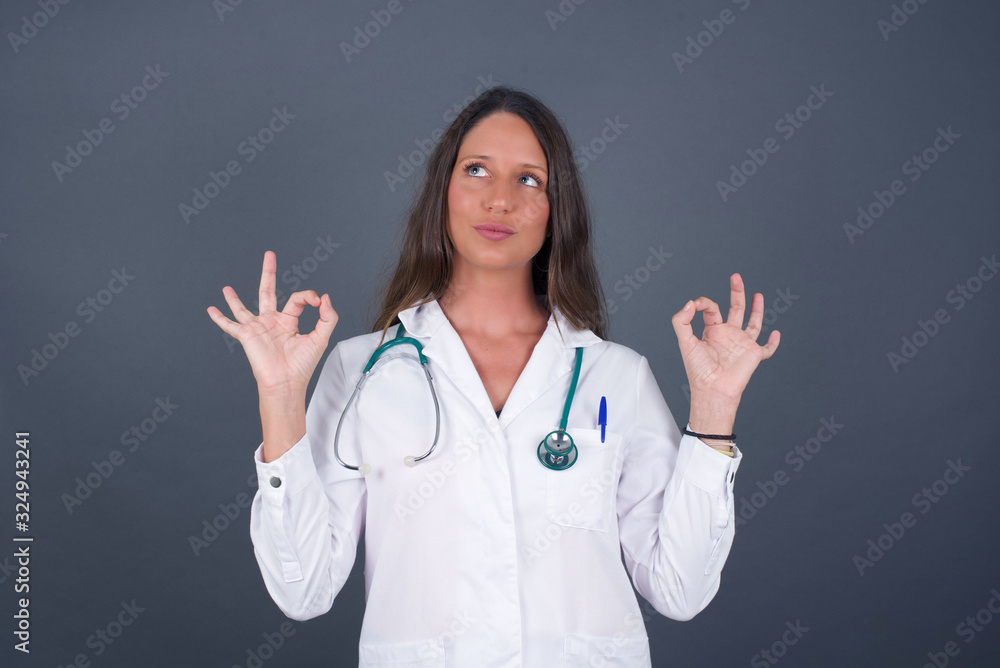 Glad attractive doctor woman shows ok sign with both hands as expresses approval, has cheerful expression being optimistic. Standing against gray wall. Glad attractive doctor woman shows ok sign with both hands as expresses approval, has cheerful expression being optimistic. Standing against gray wall.