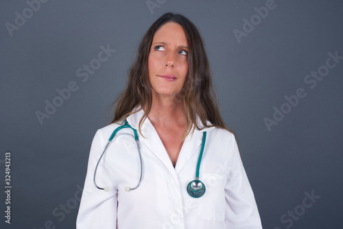 Photo of amazed puzzled young caucasian doctor female curves lips and has worried look  sees something awful in front  isolated on white background  wearing medical uniform.
