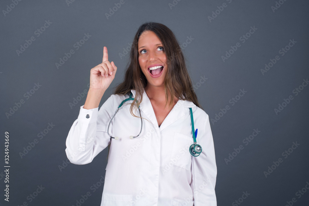 Close-up portrait of charming young doctor woman in medical uniform pointing with one finger up, looking up and smiling, I have an idea, I know the answer or the solution. Close-up portrait of charming young doctor woman in medical uniform pointing with one finger up, looking up and smiling, I have an idea, I know the answer or the solution.
