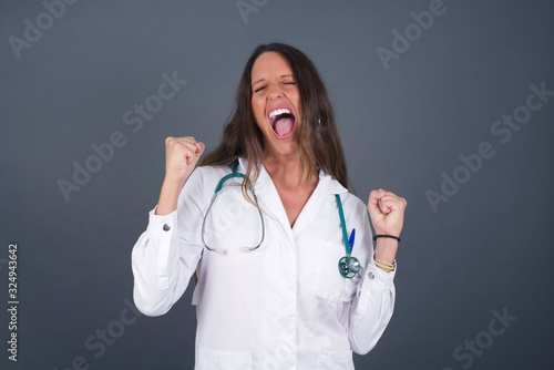 Beautiful young doctor woman wearing medical uniform being happy and excited expressing winning gesture. Successful and celebrating victory  triumphant  gray background.