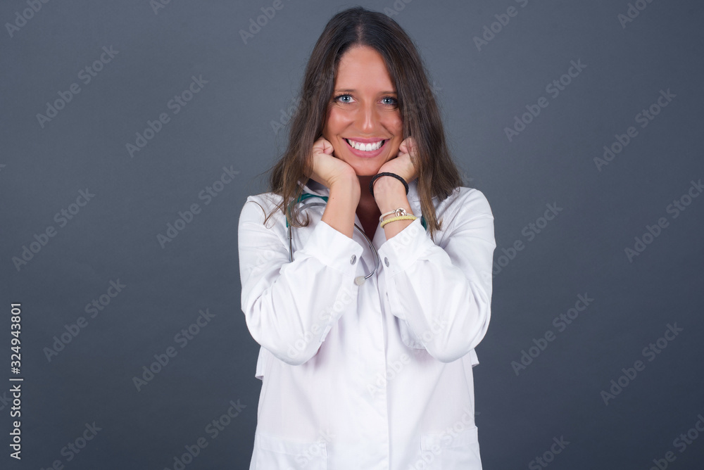 Dreamy young European woman keeps hands pressed together under chin, looks with happy expression, has toothy smile, isolated over gray background Dreamy young European woman keeps hands pressed together under chin, looks with happy expression, has toothy smile, isolated over gray background