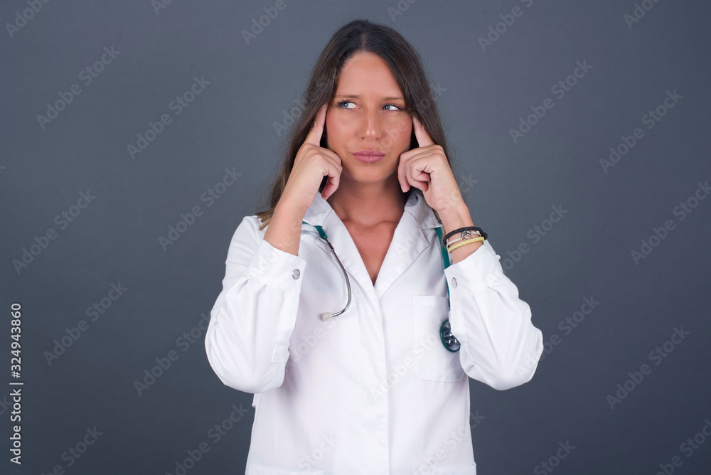 Dreamy Caucasian doctor female with thoughtful expression, looks away, keeps hand near face, thinks about something pleasant, poses against gray wall. Dreamy Caucasian doctor female with thoughtful expression, looks away, keeps hand near face, thinks about something pleasant, poses against gray wall.