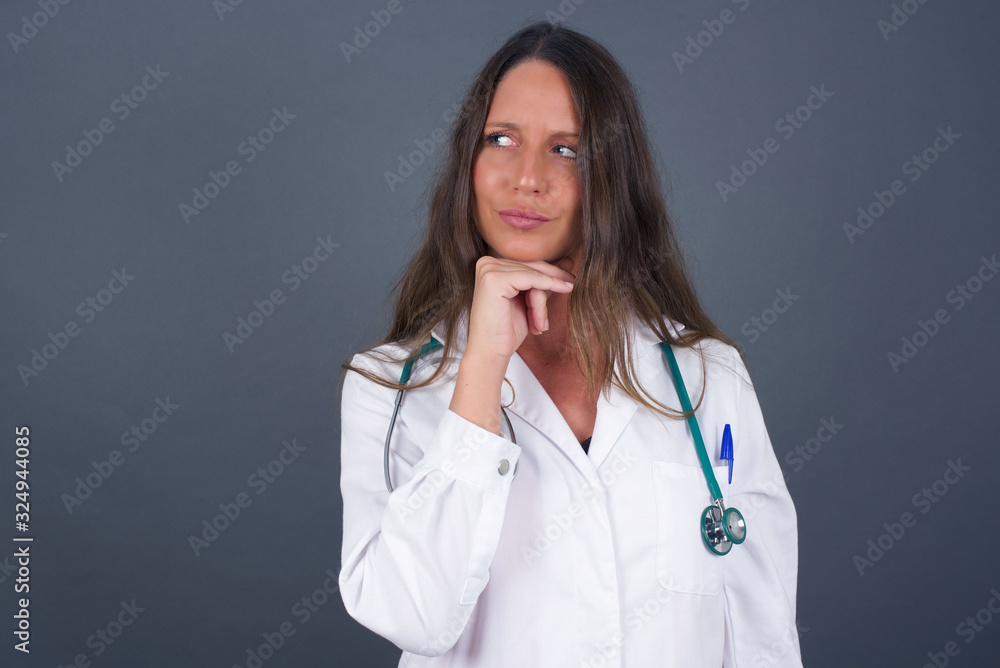 Dreamy doctor female with pleasant expression, wearing medical uniform, looks sideways, keeps hand under chin, thinks about something pleasant, poses against gray background.