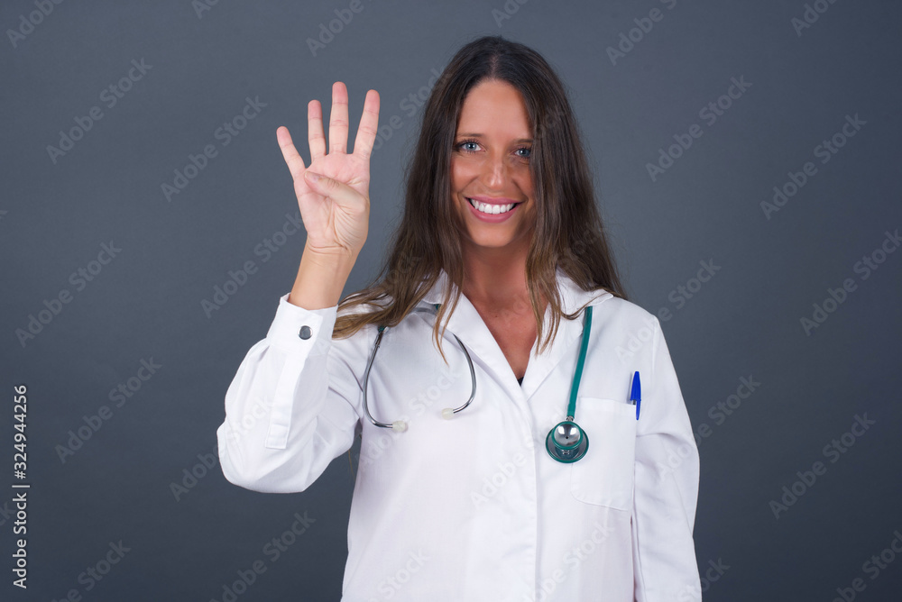 Young doctor woman standing against gray wall showing and pointing up with fingers number four while smiling confident and happy.