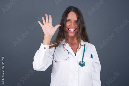 Young doctor woman standing against gray wall showing and pointing up with fingers number five while smiling confident and happy.