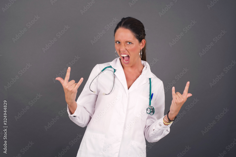 Born to rock this world. Joyful doctor woman in medical uniform screaming out loud and showing with raised arms horns or rock gesture, expressing excitement of being on concert of band. Born to rock this world. Joyful doctor woman in medical uniform screaming out loud and showing with raised arms horns or rock gesture, expressing excitement of being on concert of band.
