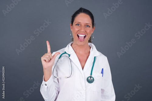 Close up portrait of pleasant looking blue eyed young doctor woman model has clever expression  raises one finger  remembers herself not to forget tell important thing. Indoors
