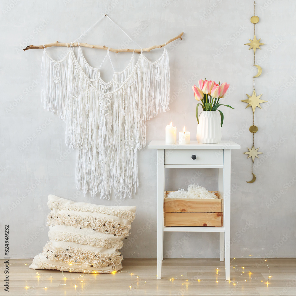 Spring boho home interior decor: macrame wall hanging decoration, white  bedside table, vase, pink tulips flowers, lighted candles, pillow, led  garland glowing lights. Light cozy modern stylish room. foto de Stock