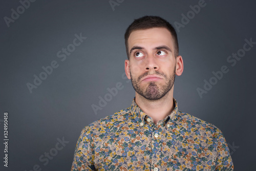 Troublesome puzzled Caucasian man frowns face, bites lip, raises eyebrows, looks up, has problems, dressed in fashionable clothes, isolated over gray background, has regret look.