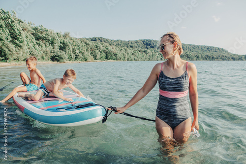 Caucasian woman parent riding kids children boys on paddle sup surfboard in water. Modern outdoor summer fun family activity. Individual aquatic seasonal sport hobby. Healthy lifestyle.