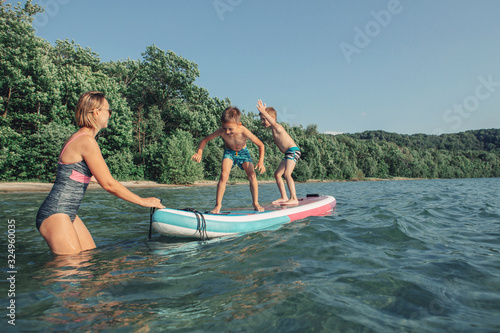 Caucasian woman parent riding kids children boys on paddle sup surfboard in water. Modern outdoor summer fun family activity. Individual aquatic seasonal sport hobby. Healthy lifestyle.