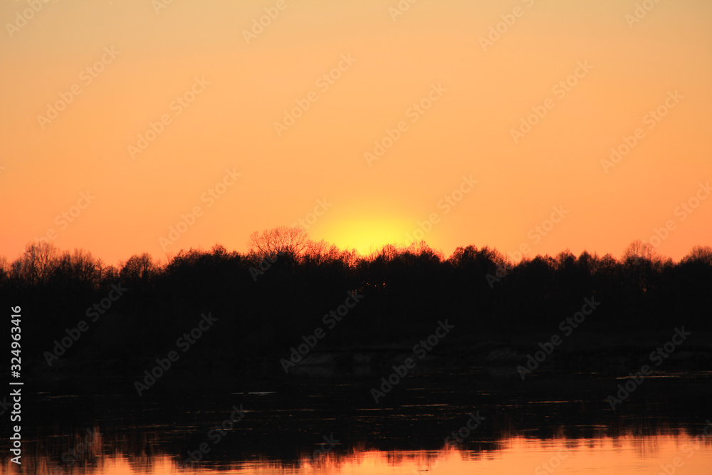 orange sunset behind the forest over the river in early spring