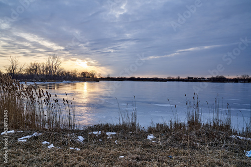 Scenic View of Winter Lake Sunset with Blue Sky and Iced Over Lake with Cattails and Trees 