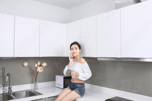 Young beautiful Asian woman talking by smartphone and drinking coffee while sitting in kitchen.