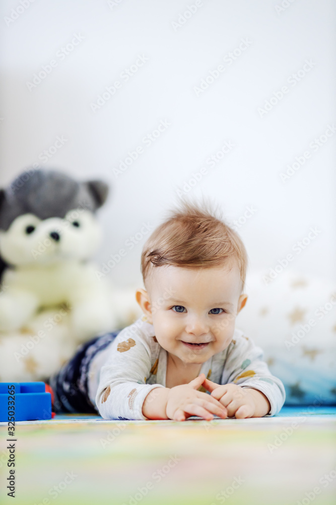 Adorable smiling blond toddler lying on stomach on the floor and crawling towards camera while looking at it. Adorable smiling blond toddler lying on stomach on the floor and crawling towards camera while looking at it.