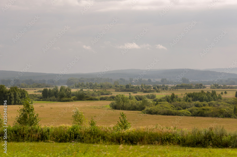 Calm blue sky in the early autumn sky over green fields, trees, forests and huge mountains. A lot of meadow herbs around. Day. Travel through nature landscape