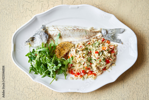 Grilled sea bass. Salad and rice with fresh vegetables. Serving on a white plate on wooden table. Restaurant menu.