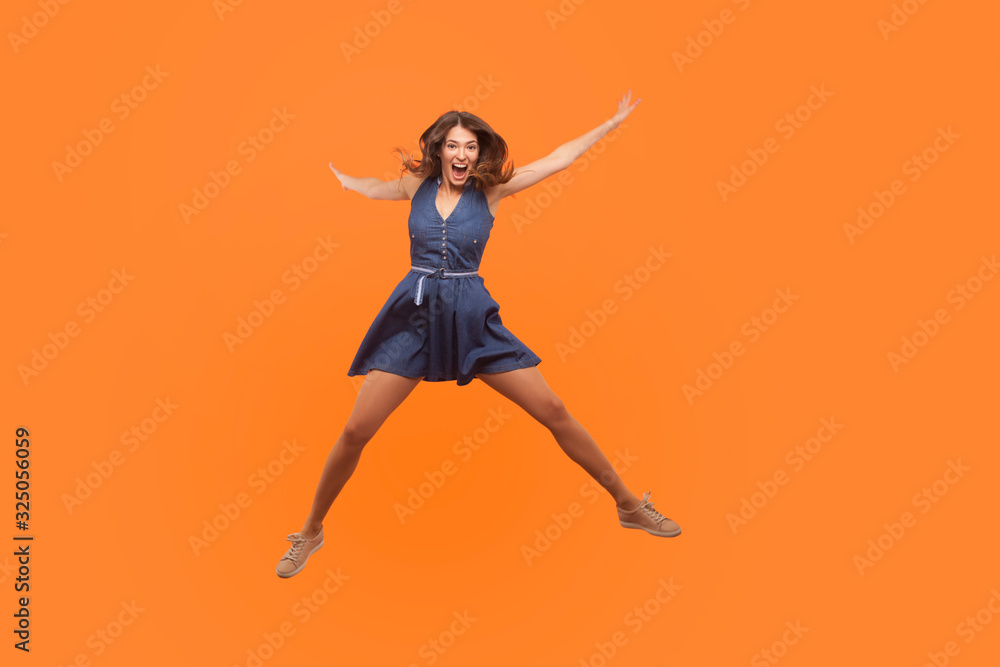 Overjoyed excited ecstatic brunette woman in denim dress jumping up like star and shouting from enthusiasm, flying isolated on orange background, full of happy joyful emotions. Freedom and energy