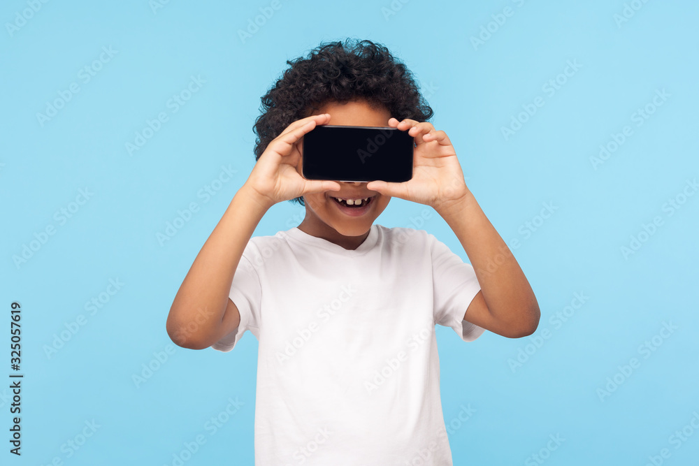 Positive lovely happy little boy with curly hair covering eyes with cellphone and smiling, unknown child hiding face with mobile phone, anonymous user. indoor studio shot isolated on blue background Positive lovely happy little boy with curly hair covering eyes with cellphone and smiling, unknown child hiding face with mobile phone, anonymous user. indoor studio shot isolated on blue background