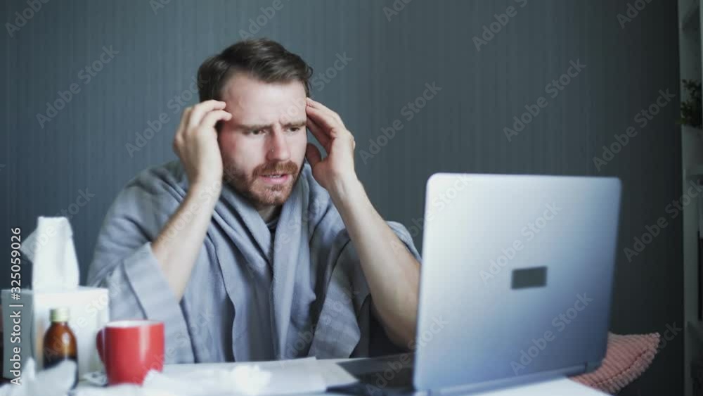 Sick business man working with laptop computer and documents at home. Sick man is suffering from severe headache touching his head massaging temples indoors at home. Sick business man working with laptop computer and documents at home. Sick man is suffering from severe headache touching his head massaging temples indoors at home.